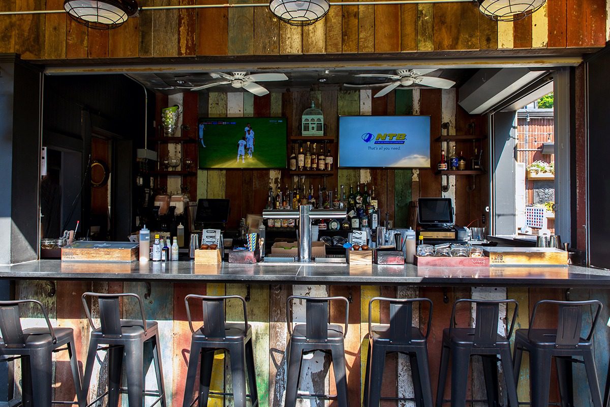 Modern bar counter with multiple stools and two TVs mounted above it.
