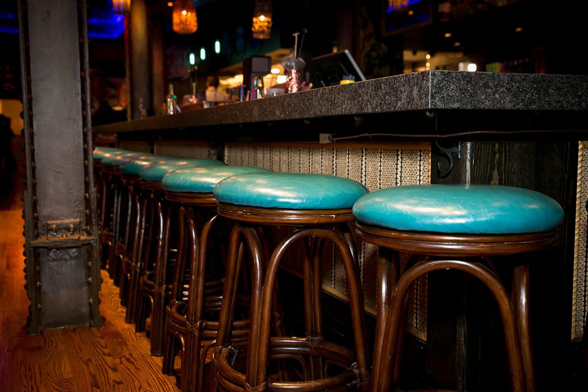 A row of teal cushioned bar stools at a dimly lit bar counter.