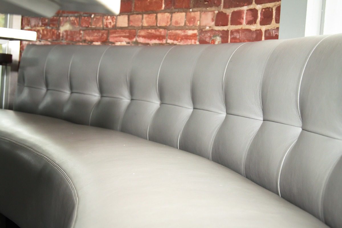 Curved gray leather sofa with tufted backrest against a brick wall.