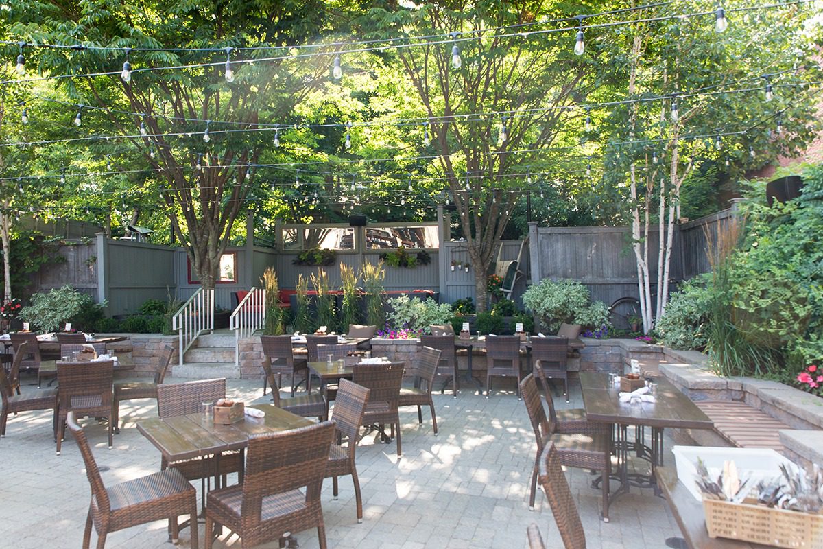 Outdoor dining area with wooden tables and chairs under leafy trees.