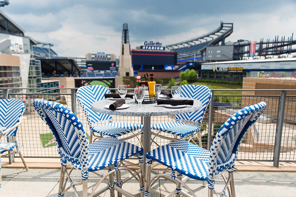 Outdoor stadium seating with blue and white chairs around a table, overlooking a sports field.