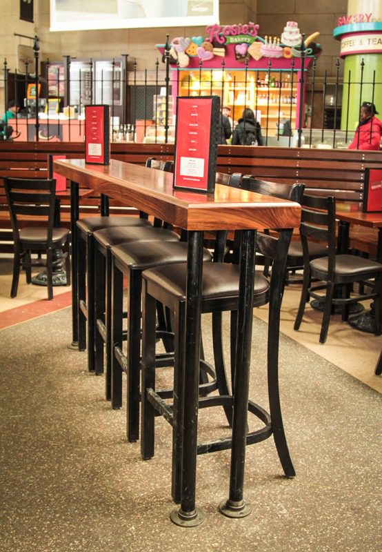 Tall wooden stools lined up along a high wooden table in a cozy restaurant.